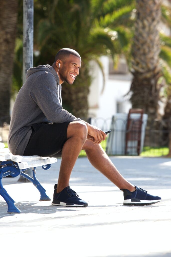 Full body portrait of fit black guy sitting on bench and listening music